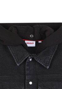 Zwarte denimjas met een knoopsluiting aan de voorkant, klassieke kraag en capuchon. Heeft zilverkleurige drukknoopsluitingen en een HUGO-label in rood en wit.