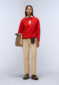 Κόκκινο φούτερ με σχέδιο, συνδυασμένο με πράσινο πουκάμισο με καρό, μπεζ παντελόνια και καφέ παπούτσια. Κρατάει καφέ τσάντα και αξεσουάρ.