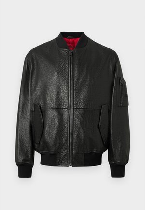 Černá kožená bomber bunda s texturovaným povrchem, zapínáním na zip, žebernatým pasem a bočními kapsami. Má červenou vnitřní podšívku a pracovní kapsu.