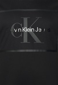 Czarna bluza Calvin Klein Jeans o gładkiej fakturze z dużym wypukłym logo CK i panelem z siateczki, podkreślająca nazwę marki.