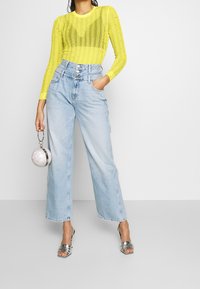 Gul långärmad topp med ribbad textur och utsmyckningar, kombinerad med högmidjade ljust blå vida jeans och silverfärgade klacksandaler.