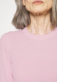 Äldre kvinna med axellångt grått hår som bär en ljusrosa, strukturerad tröja, sedd från axlarna till läpparna mot en enkel bakgrund.