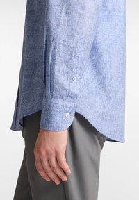 Camisa de manga larga azul claro con un patrón, puños abotonados y un dobladillo curvado, combinada con pantalones grises. El material parece texturizado.
