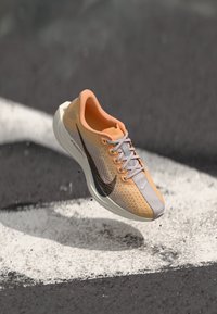 Pantof de alergare Nike de culoare portocalie deschis și gri, cu swoosh negru, în aer deasupra asfaltului cu marcaje rutiere albe.