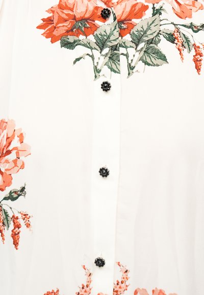 Λευκό ύφασμα με μεγάλα πορτοκαλί λουλουδένια σχέδια και πράσινα φύλλα, διακοσμημένο με μαύρα κουμπιά σε σχήμα λουλουδιού κατά μήκος του κέντρου.