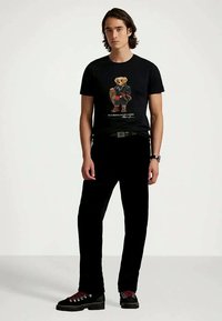 Ung man med svart grafisk t-shirt med en björn i läderjacka, svarta byxor, svart bälte, armbandsur och svarta skor med röda snören.