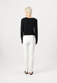 Svart langermet topp med pyntede skuldre, kombinert med hvite skinny jeans. Modellen står med ryggen til, og viser frem designet og passformen bak.