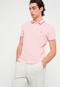 Ροζ polo μπλούζα με κοντά μανίκια και γιακά, με δύο κουμπιά, μανσέτες σε ναυτικό μπλε και λευκό ριγέ, και μικρό λογότυπο στο στήθος.