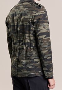 Person iført en mørkegrøn og sort zigzag-mønstret militærjakke, vist fra ryggen mod en ensfarvet baggrund.