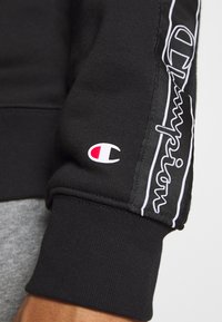 Felpa nera con logo "Champion" ricamato in bianco sul braccio e un piccolo emblema rosso e bianco. Tessuto morbido con polsini a coste.