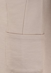 Beiges Gewebe mit einer aufgenähten quadratischen Tasche, die die detaillierte Naht und die Textur des Materials zeigt.