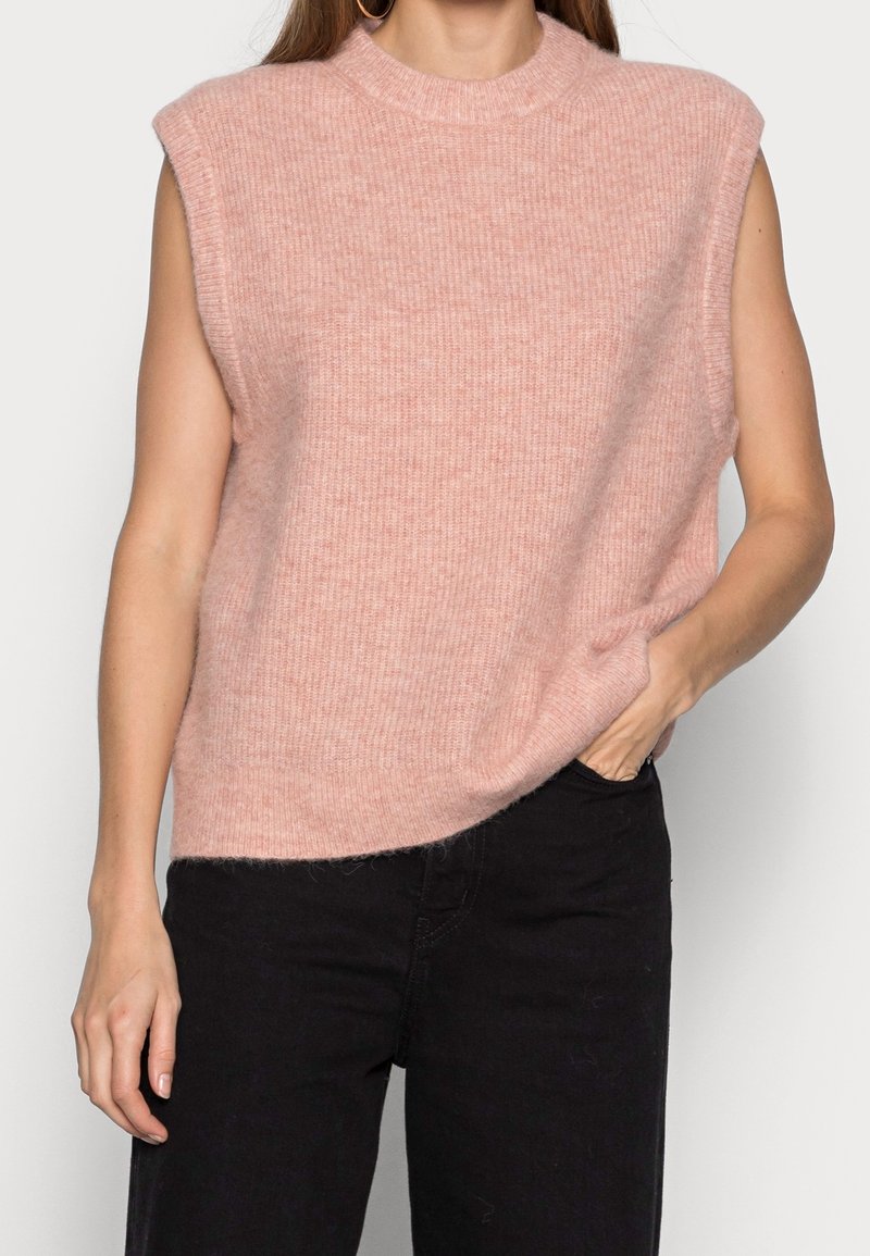 Bezramienny, jasnoróżowy, dziergany sweter z wysokim kołnierzem i subtelną fakturą, zestawiony z czarnymi dżinsami, ręka spoczywająca po boku.
