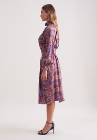 Lila Kleid mit Paisleymuster, langen Ärmeln, Kragen und tailliertem Schnitt, kombiniert mit schwarzen High-Heels.