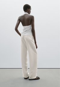 Bílý halter top s volánkovým okrajem, doplněný volnými, krémově bílými kalhotami s širokými nohavicemi. Model stojí na šedé podlaze před světlým pozadím.