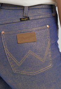 Plave traperice s Wrangler logom, zlatim šavovima i bakarnim zakovicama, s karakterističnim cik-cak dizajnom na stražnjem džepu.