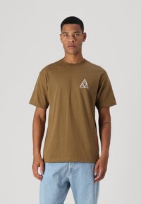 Camiseta de algodón marrón con mangas cortas y escote redondo. Presenta un gráfico triangular blanco en el lado izquierdo del pecho. Se lleva con pantalones vaqueros de color azul claro.