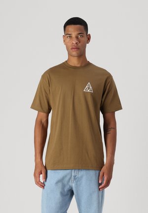 Camiseta de algodón marrón con mangas cortas y escote redondo. Presenta un gráfico triangular blanco en el lado izquierdo del pecho. Se lleva con pantalones vaqueros de color azul claro.