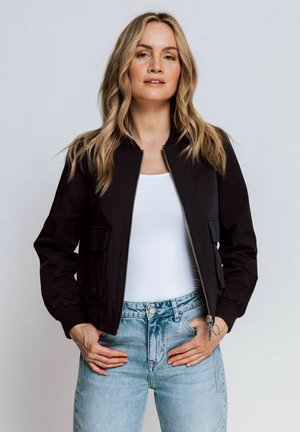Black bomber jacket with zip front, ribbed cuffs, and dual side pockets. Paired with a white tank top and light blue denim jeans.