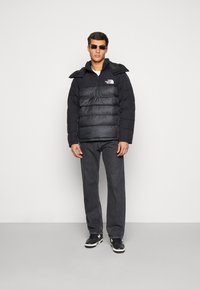 Giacca a vento nera con zip frontale, caratterizzata da pannelli orizzontali e un logo, abbinata a jeans scuri e sneakers nere.