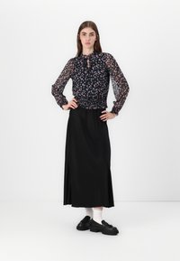 Gonna nera maxi con stampa floreale, blusa trasparente a maniche lunghe, vita smock e dettagli con bottoni. Indossata con scarpe nere e calzini bianchi.