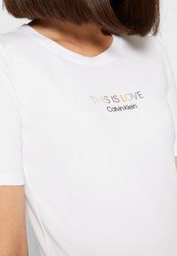 Žena v bílém tričku Calvin Klein s textem "TOTO JE LÁSKA" v duhových barvách na hrudi.