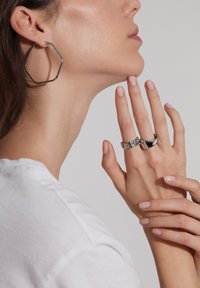 Sølv hexagonale hoop øreringe, en sølv chunky ring og en glat, buet sølvring på en hånd med neutral neglelak, mod en lys baggrund.