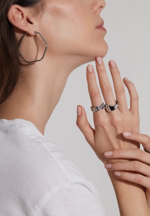 Sølv hexagonale hoop øreringe, en sølv chunky ring og en glat, buet sølvring på en hånd med neutral neglelak, mod en lys baggrund.