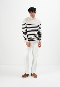 Uomo che indossa un maglione a collo alto a righe crema e blu navy, pantaloni bianchi e scarpe ciaspole marroni, appoggiato su uno sfondo bianco uniforme.