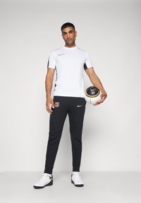 Nike sportoutfit bestaande uit een witte, korte mouw top met zwarte accenten en zwarte trainingsbroek met gouden logo-details. Houdt een voetbal vast.