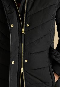 Must muljetud jakk kahe kuldse tõmbetaja, kuldsete nööpide ja küljetaskutega, seljas inimesel, kelle käed on taskutes.