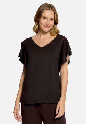 Femme aux cheveux blonds mi-longs portant un chemisier noir à manches courtes avec des détails en dentelle et un pantalon noir assorti, souriant légèrement.