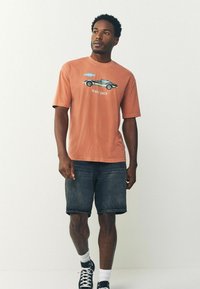 Orange bomulds T-shirt med korte ærmer og rund hals, med et trykt grafisk motiv af en bil og teksten "Hold tempo."