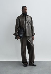 Hnedá kožená košela s vysokým golierom a horizontálnym šitím, spárovaná s voľnými hnedými koženými nohavicami. Drží čiernu koženú tašku.