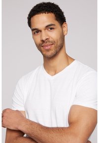 Una camiseta blanca de cuello en V en un hombre con cabello oscuro y rizado. La camiseta tiene una textura suave y un diseño ajustado, que la lleva con los brazos cruzados.