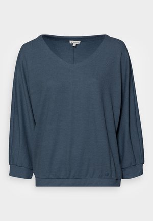 Haut à col en V, coupe ample en tissu texturé bleu foncé. Présente des manches longues et aérées ainsi qu'une ourlet incurvé avec un accent de logo en bas.