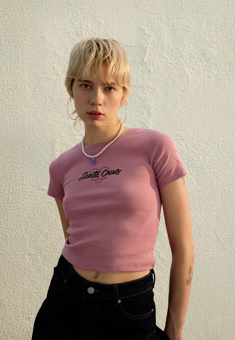 Ružičasta kratka majica s crnim logotipom "Santa Cruz" i kratkim rukavima, uparena s crnim trapericama s visokim strukom. U pozadini se nalazi teksturirani bijeli zid.