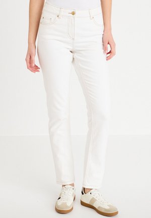 Kobieta ubrana w białe jeansy o prostych nogawkach i beżowo-białe trampki, stojąca na tle jednolitego białego tła.