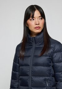 Giacca puffer blu navy con colletto alto, design trapuntato e tasche con zip. Realizzata in materiale sintetico liscio con una silhouette aderente.