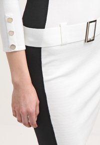 Robe en maille composée de sections de tissu blanc et noir, avec une large ceinture blanche et une boucle métallique rectangulaire ; détails de boutons argentés sur les manches.