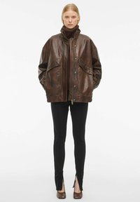 Pruun nahktagi suurte küljetaskutega, kõrge kraega, koos mustade liibuvate pükstega, millel on lõhikud, ja pruunide kõrgete kontsadega kingadega.