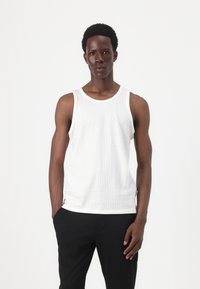 Hvit ribbet tanktop med rund halslinje og brede skulderstropper, kombinert med svarte bukser. Stoffet har en myk, teksturert overflate.