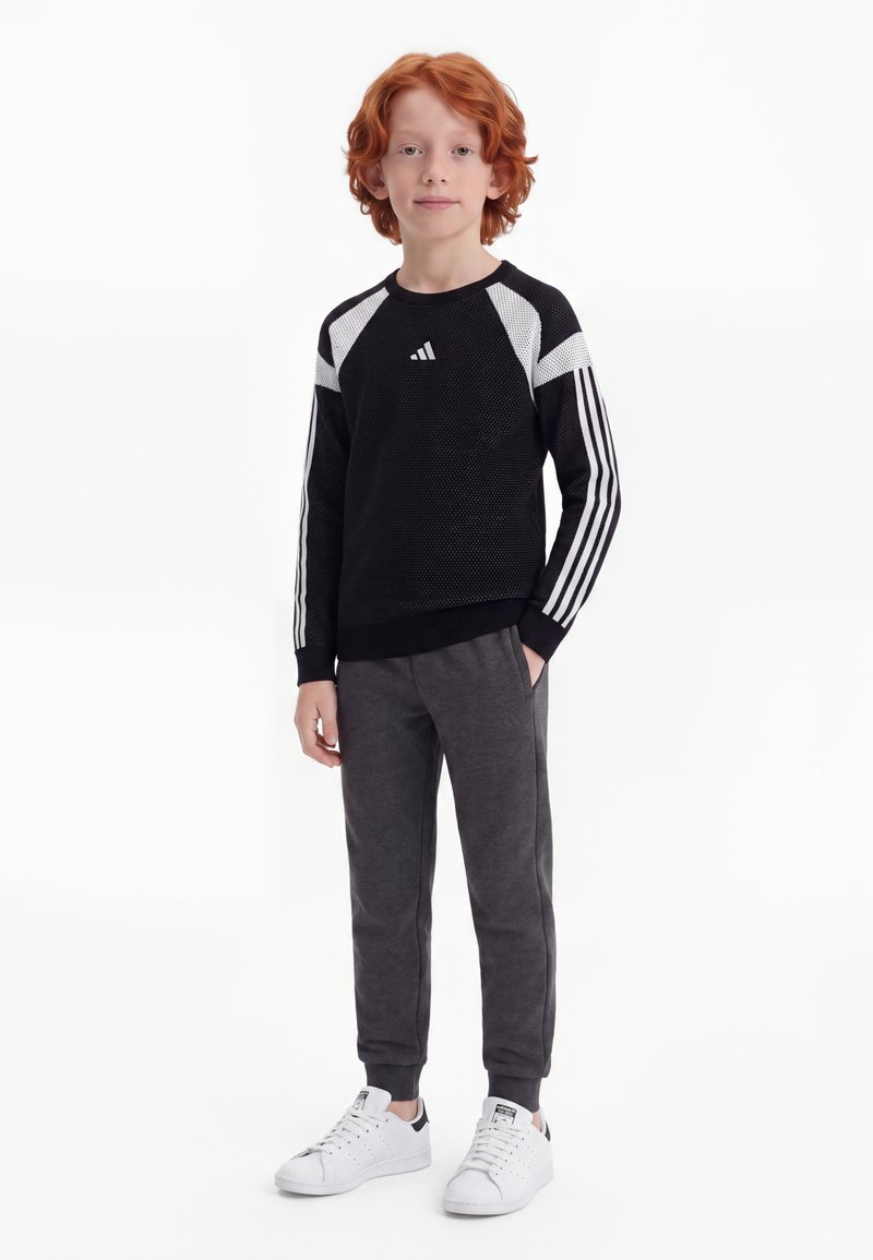 Enfant aux cheveux roux bouclés portant un pull de sport noir et blanc, un pantalon de jogging gris foncé et des baskets blanches, debout sur un fond blanc.