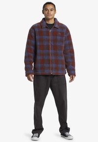 Veste en polaire à carreaux bruns et bleus, avec fermeture éclair sur le devant, grand col et coupe ample. Portée avec un pantalon sombre et des baskets noires.