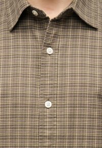 Καφέ και μπεζ πουκάμισο με καρό μοτίβο και λευκά κουμπιά, μερικώς κουμπωμένο στον γιακά στο πάνω μέρος του στήθους ενός ατόμου.