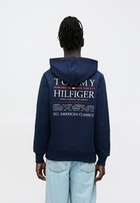 Άτομο με ντραντλοκς που φοράει μπλε σκουρόχρωμη φούτερ με κουκούλα με το λογότυπο Tommy Hilfiger και ανοιχτόχρωμα τζιν, με την πλάτη προς το μέρος μας, απέναντι σε απλό φόντο.