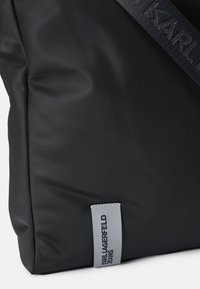 Borsa tote in nylon nero caratterizzata da una texture liscia e un'etichetta grigia con il logo "KARL LAGERFELD JEANS", completata da una robusta tracolla grigia.