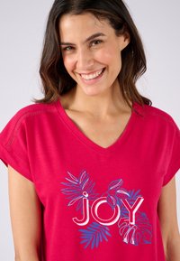 Camiseta rosa de cuello en V con un diseño floral y la palabra "ALEGRÍA" en contornos blancos bold, mangas cortas y hombros texturizados. Tela suave.