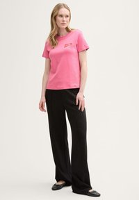 Rosa bomullströja med kort ärm och en liten röd logotyp, i kombination med svarta vida byxor och svarta flats.
