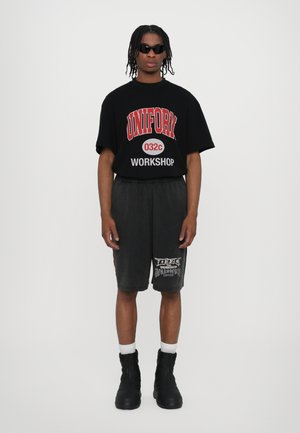 Schwarzes, oversized T-Shirt mit rotem Text und Grafiken, kombiniert mit grauen Shorts mit weißem Aufdruck. Schwarze Stiefel und weiße Socken runden den Look ab.