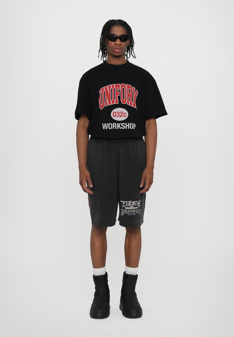 Black oversized T-shirt with red text and graphics, paired with grey shorts featuring white print. Black boots and white socks complete the look.